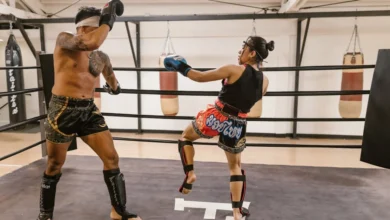 Dois lutadores praticando Muay Thai em um ringue com sacos de pancada ao fundo.