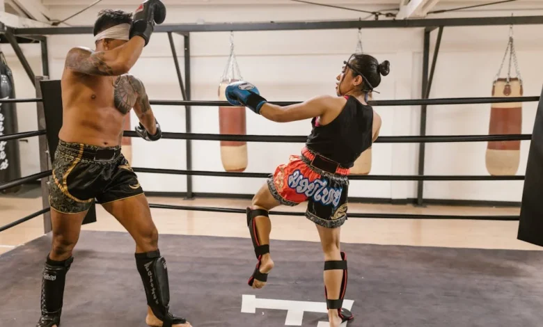 Dois lutadores praticando Muay Thai em um ringue com sacos de pancada ao fundo.